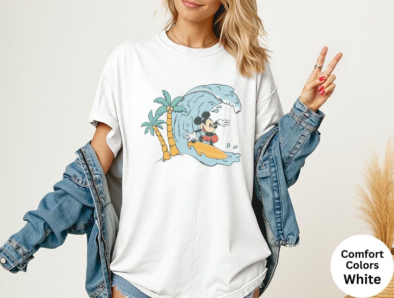 May include: White t-shirt featuring a cartoon Mickey Mouse surfing a wave. The design includes palm trees and a yellow surfboard. The shirt has the words "Comfort Colors White" on the bottom right.