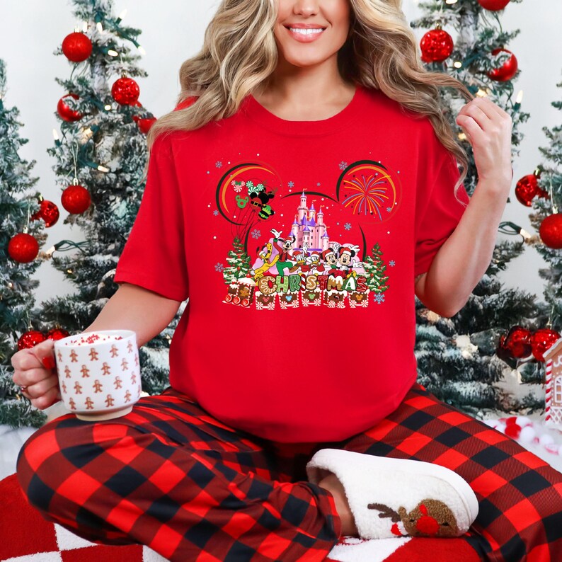 May include: A red t-shirt featuring a Disney Christmas design with Mickey Mouse ears, a castle, and various characters. The shirt is paired with red and black plaid pajama pants and a mug with gingerbread men. Christmas tree decorations are in the background.
