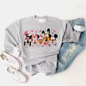 Op de afbeelding: Een grijs sweatshirt met een grafische print van Mickey Mouse, Minnie Mouse, Donald Duck, Goofy en Pluto die roze hartvormige zonnebrillen dragen. De print bevat ook harten, sterren en het woord "Love!" in roze.