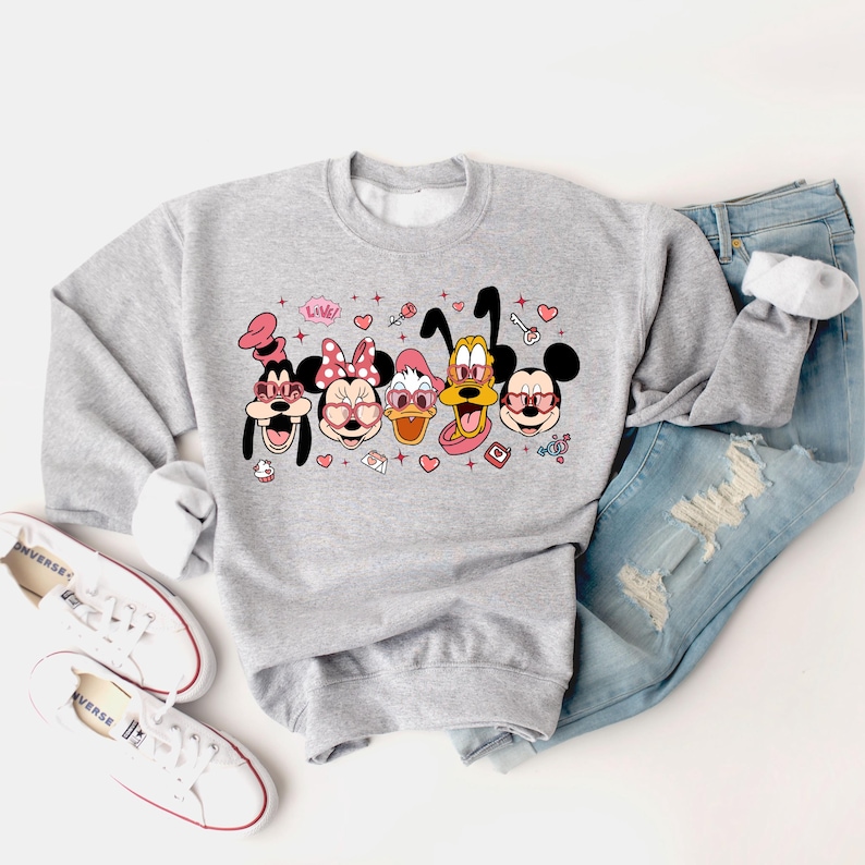 Op de afbeelding: Een grijs sweatshirt met een grafische print van Mickey Mouse, Minnie Mouse, Donald Duck, Goofy en Pluto die roze hartvormige zonnebrillen dragen. De print bevat ook harten, sterren en het woord "Love!" in roze.