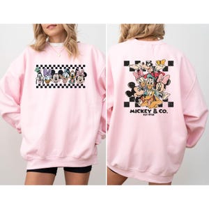 May include: Pink sweatshirt featuring a graphic of Disney characters on a black and white checkered background. The back of the sweatshirt displays a group of Disney characters and the text "Mickey & Co. EST 1928".