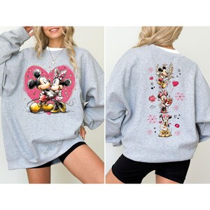 May include: A gray sweatshirt featuring a large pink heart with Mickey and Minnie Mouse embracing. The back of the sweatshirt has a vertical design with Mickey, Minnie, Pluto, and hearts. The sweatshirt has a white collar.