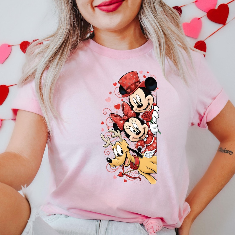 Puede incluir: Camiseta rosa claro con un dise&ntilde;o de San Valent&iacute;n con Mickey Mouse, Minnie Mouse y Pluto. Mickey lleva un sombrero de copa, Minnie un lazo y Pluto un collar con forma de coraz&oacute;n. El dise&ntilde;o incluye corazones y una rosa.