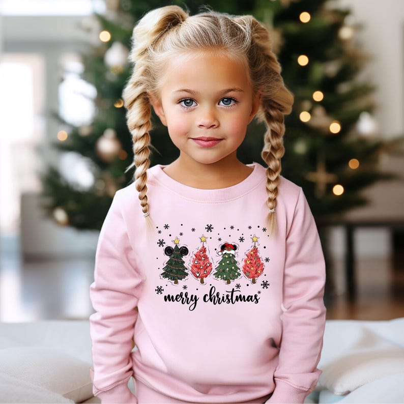 May include: A young girl wearing a light pink sweatshirt with a Christmas tree design. The design features four colorful Christmas trees with stars and the words "merry christmas". The background includes a Christmas tree with lights.