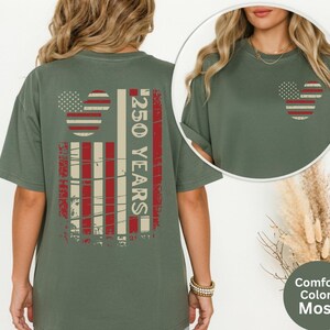 May include: A moss green t-shirt with a distressed American flag design on the back and a smaller version on the front. The back design includes the text "250 YEARS" in white. The shirt is a Comfort Colors brand.