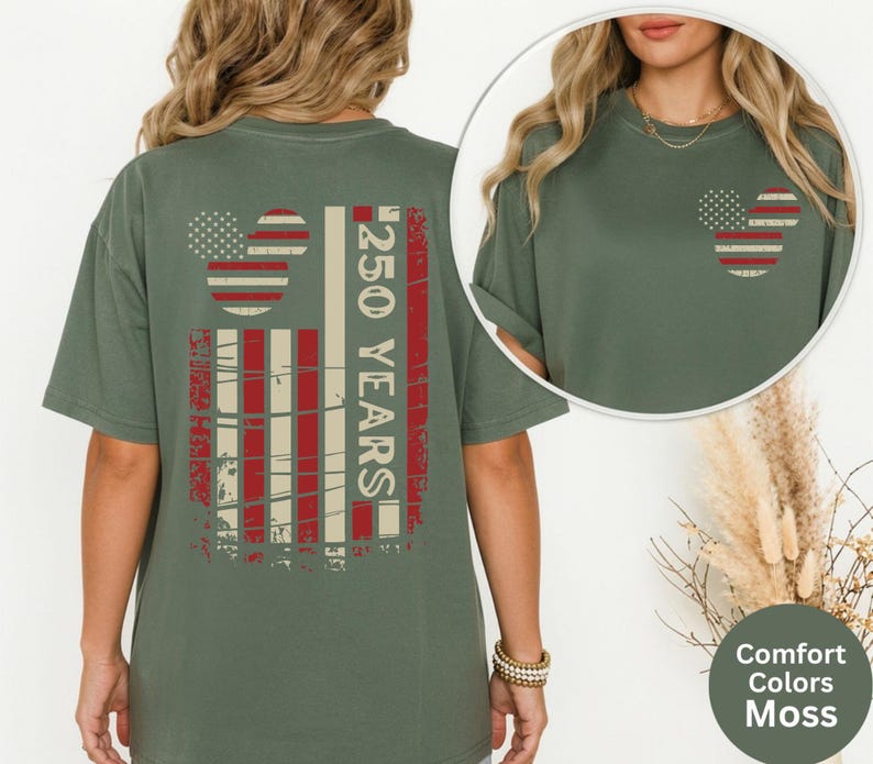May include: A moss green t-shirt with a distressed American flag design on the back and a smaller version on the front. The back design includes the text "250 YEARS" in white. The shirt is a Comfort Colors brand.
