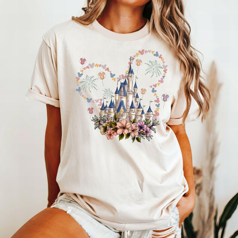 May include: Cream-colored t-shirt featuring a whimsical castle design with blue turrets, surrounded by floral accents and a heart-shaped arrangement of balloons and fireworks. The design is centered on the front of the shirt.