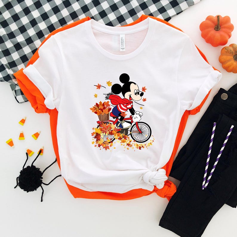 May include: White t-shirt featuring a cartoon Mickey Mouse riding a red bicycle through autumn leaves. Mickey wears a red and white striped scarf and a basket of leaves. Black pants and pumpkins are in the background.