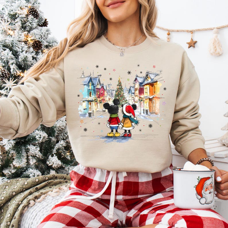 Op de afbeelding: Beige sweatshirt met een aquarel illustratie van Mickey en Minnie Mouse in een winterse sc&egrave;ne. Het ontwerp bevat een kerstboom, kleurrijke gebouwen en vallende sneeuwvlokken. De sweatshirt wordt gecombineerd met een rood-witte geruite pyjamabroek.