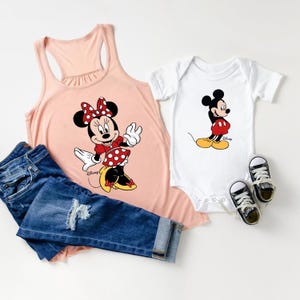 K&ouml;nnte beinhalten: Ein rosafarbenes Tanktop mit einem Minnie Mouse-Motiv und ein wei&szlig;er Body mit einem Mickey Mouse-Motiv. Das Tanktop hat einen R&uuml;schenkragen und der Body hat einen Druckknopfverschluss. Das Tanktop wird mit einer blauen Jeans kombiniert.