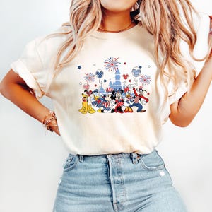 K&ouml;nnte beinhalten: Cremefarbenes T-Shirt mit einem patriotischen Disney-Design. Die Grafik zeigt Micky und Minnie Maus, Donald Duck, Goofy und Pluto, zusammen mit Feuerwerk, Luftballons und einem Schloss. Die Charaktere sind in Rot, Wei&szlig; und Blau gekleidet.