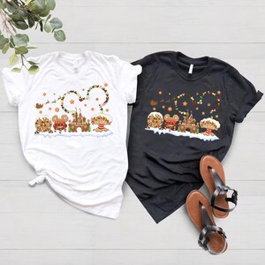 May include: Two t-shirts featuring a Christmas gingerbread house and Mickey Mouse characters. The white t-shirt has a red and green garland with stars. The black t-shirt has a red and green garland with stars. Both shirts feature a gingerbread house and Mickey Mouse characters.