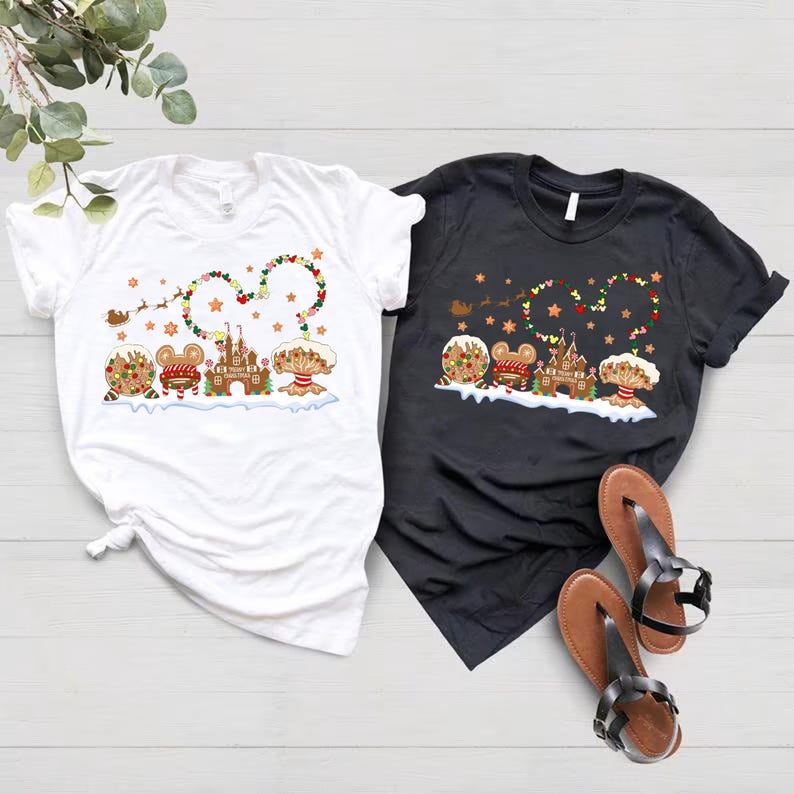 May include: Two t-shirts featuring a Christmas gingerbread house and Mickey Mouse characters. The white t-shirt has a red and green garland with stars. The black t-shirt has a red and green garland with stars. Both shirts feature a gingerbread house and Mickey Mouse characters.