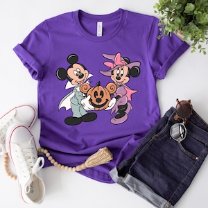 May include: Purple t-shirt with a graphic of Mickey Mouse and Minnie Mouse dressed in Halloween costumes. Mickey is a vampire and Minnie is a witch. They are holding a jack-o-lantern.