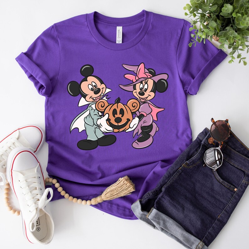May include: Purple t-shirt with a graphic of Mickey Mouse and Minnie Mouse dressed in Halloween costumes. Mickey is a vampire and Minnie is a witch. They are holding a jack-o-lantern.
