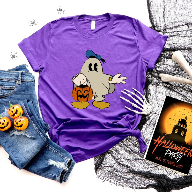 Puede incluir: Camiseta morada con un fantasma de dibujos animados que lleva un gorro de marinero y sostiene una calabaza. El fantasma lleva zapatos amarillos. La camiseta est&aacute; sobre un fondo blanco con un par de pantalones vaqueros azules, una telara&ntilde;a negra y una invitaci&oacute;n a una fiesta de Halloween.