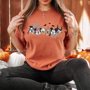 Puede incluir: Una camiseta naranja quemada con un gr&aacute;fico de Mickey Mouse, Minnie Mouse, Goofy, Donald Duck, Pluto y Mal&eacute;fica con una telara&ntilde;a y murci&eacute;lagos. Los personajes llevan todos disfraces de Halloween.