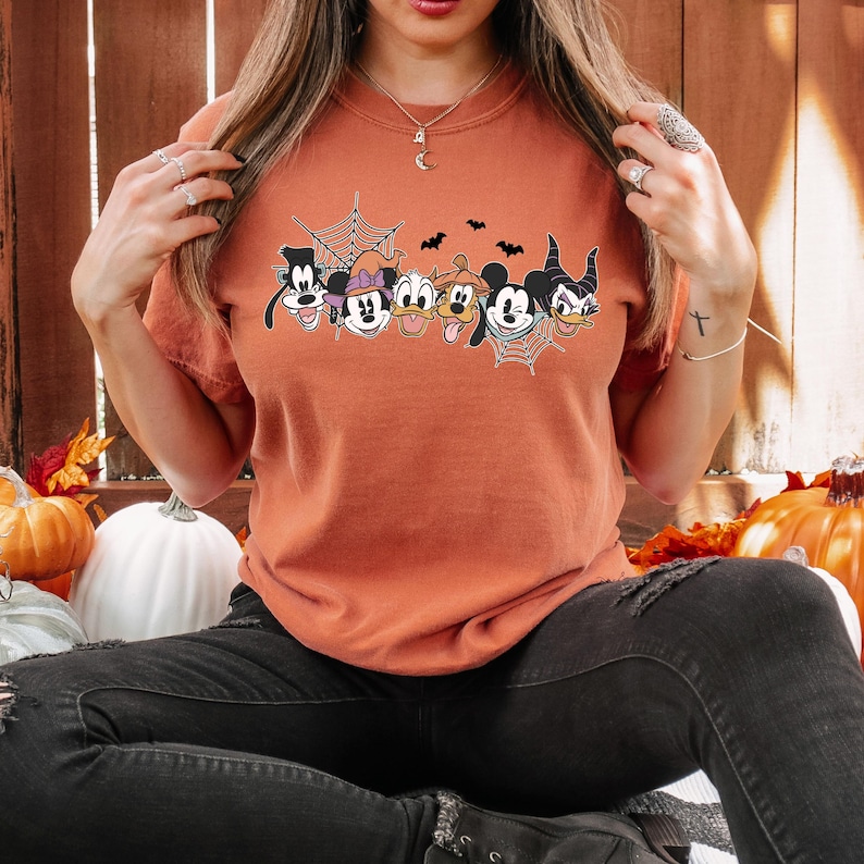 Puede incluir: Una camiseta naranja quemada con un gr&aacute;fico de Mickey Mouse, Minnie Mouse, Goofy, Donald Duck, Pluto y Mal&eacute;fica con una telara&ntilde;a y murci&eacute;lagos. Los personajes llevan todos disfraces de Halloween.