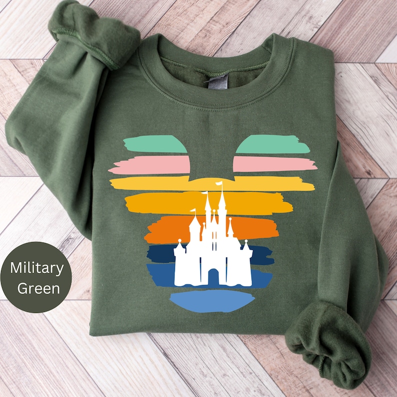 May include: A green sweatshirt with a white silhouette of Cinderella's castle inside a Mickey Mouse head shape. The Mickey Mouse head shape is made up of colorful horizontal stripes in shades of blue, green, orange, yellow, pink, and red.