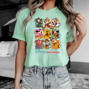 May include: A light green t-shirt featuring a colorful graphic of Disney characters in cowboy attire. The graphic includes Mickey Mouse, Minnie Mouse, Donald Duck, Daisy Duck, Chip, Dale, and Goofy. The text "Disney Howdy Partners" is printed below the graphic.