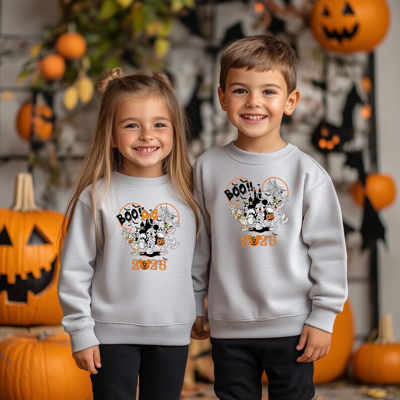May include: Two children in light grey sweatshirts featuring a Halloween design. The design includes "BOO!!", a haunted house, spiderwebs, and the year "2025". The sweatshirts are worn with black trousers.