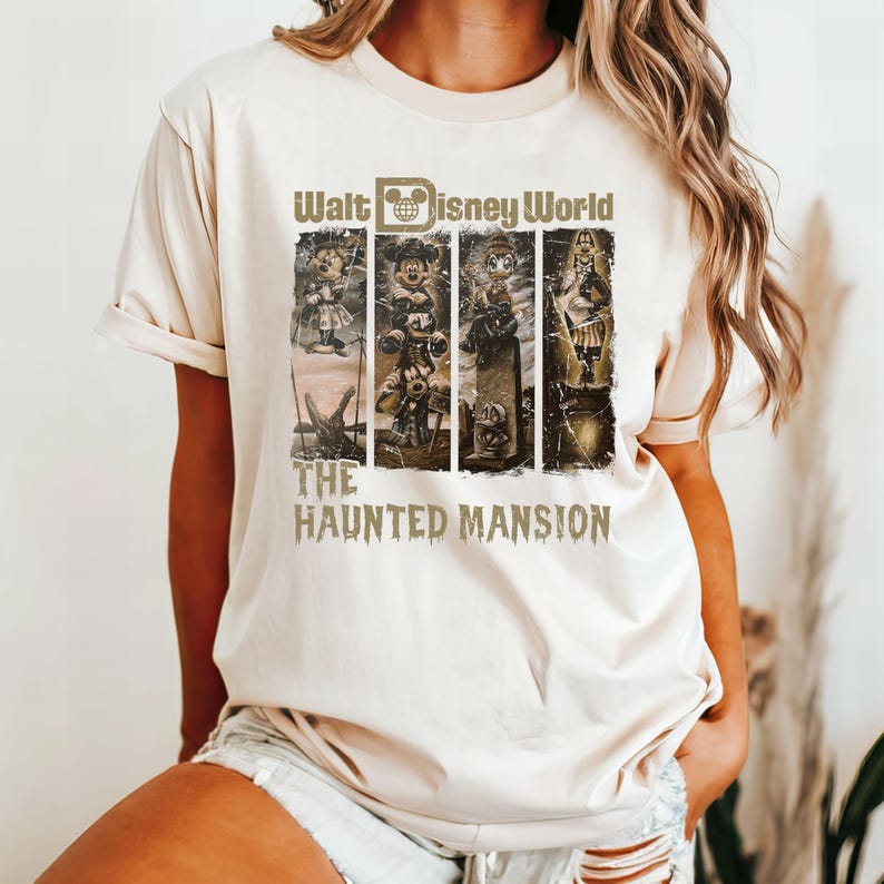 May include: A beige t-shirt featuring a vintage-style graphic of the Haunted Mansion attraction at Walt Disney World. The graphic depicts Mickey Mouse and other characters from the ride.