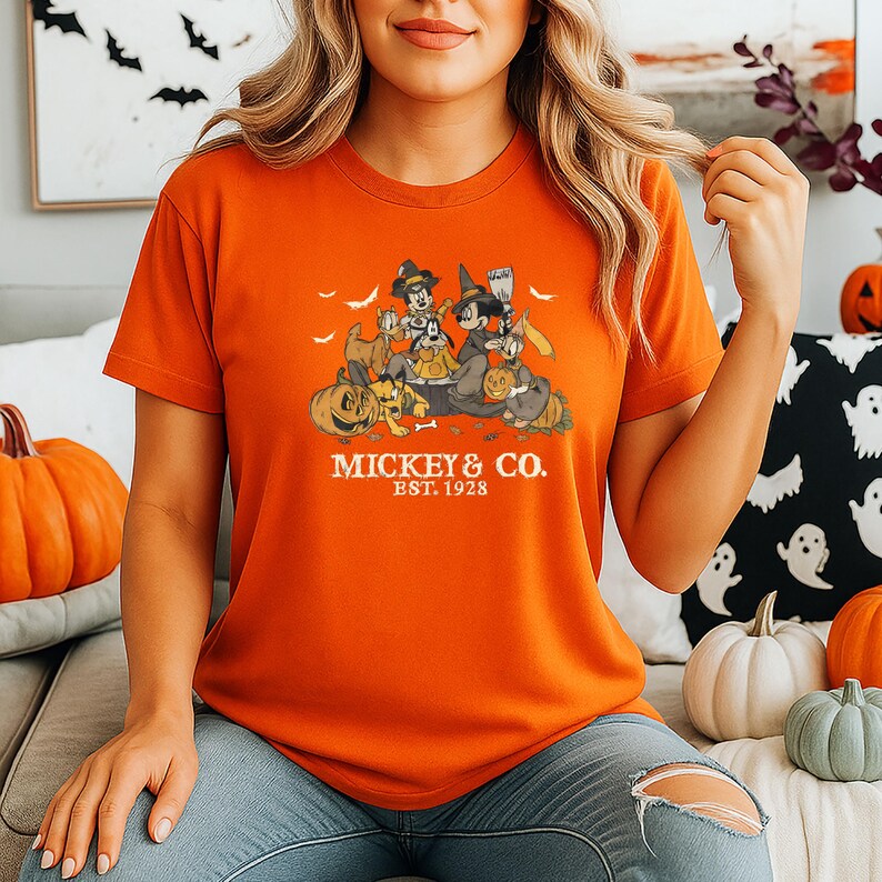 Pode incluir: Camiseta laranja com um design de Halloween com Mickey Mouse e amigos. O design inclui ab&oacute;boras, morcegos e o texto "Mickey & Co. Est. 1928". A camiseta &eacute; de gola redonda e manga curta.