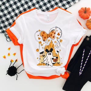May include: White t-shirt with a Halloween-themed graphic featuring a ghost holding a pumpkin, a castle, and a bow. The graphic is in orange, black, and white.