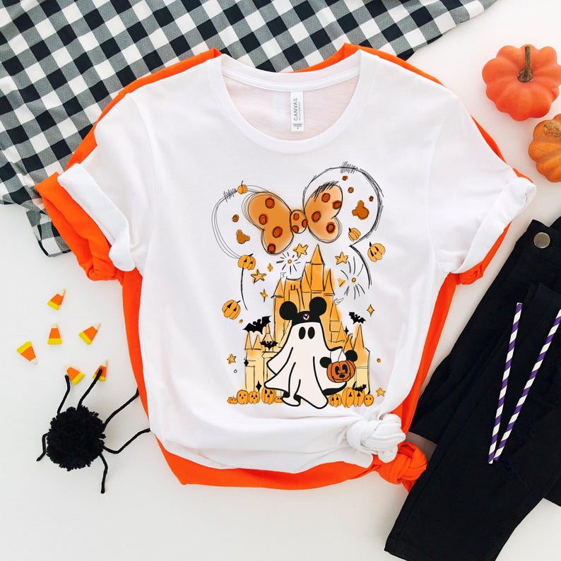 May include: White t-shirt with a Halloween-themed graphic featuring a ghost holding a pumpkin, a castle, and a bow. The graphic is in orange, black, and white.