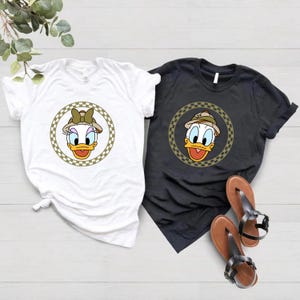 May include: Two t-shirts with a cartoon image of a duck wearing a safari hat and a bow. The duck is surrounded by a checkered circle. One shirt is white and the other is black.