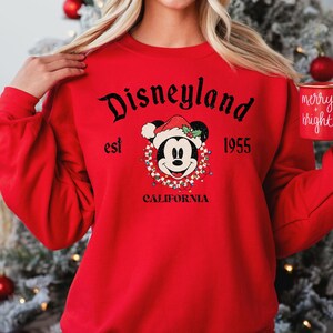 Puede incluir: Sudadera roja con un gr&aacute;fico negro de Mickey Mouse con un gorro de Pap&aacute; Noel y rodeado de luces navide&ntilde;as. El texto "Disneyland est 1955 California" est&aacute; impreso en la sudadera.