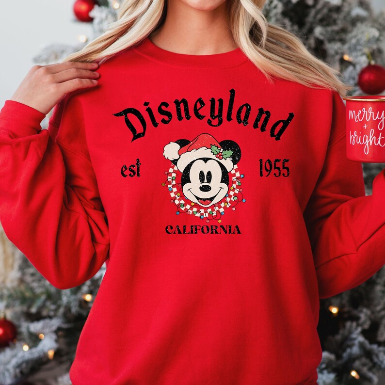 Puede incluir: Sudadera roja con un gr&aacute;fico negro de Mickey Mouse con un gorro de Pap&aacute; Noel y rodeado de luces navide&ntilde;as. El texto "Disneyland est 1955 California" est&aacute; impreso en la sudadera.