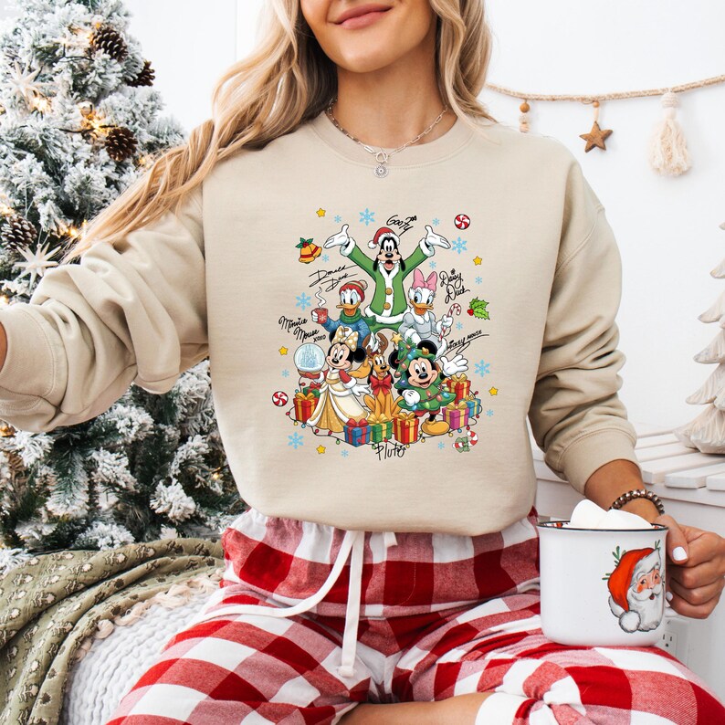 K&ouml;nnte beinhalten: Beiges Sweatshirt mit einem farbenfrohen Design von Disney-Figuren in festlicher Kleidung. Das Design umfasst Micky Maus, Minnie Maus, Donald Duck und Goofy, umgeben von Geschenken und festlichen Dekorationen. Das Sweatshirt wird mit einer rot-wei&szlig; karierten Pyjamahose kombiniert.