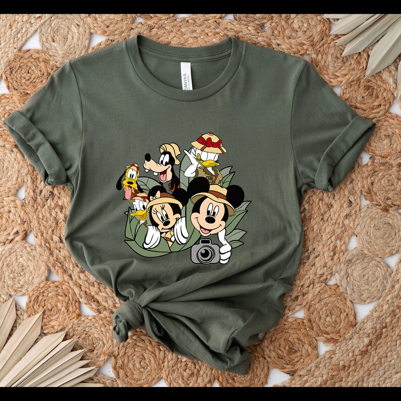 May include: A green t-shirt featuring a graphic print of Mickey Mouse, Minnie Mouse, Goofy, Donald Duck and Pluto dressed in safari gear. The characters are surrounded by green leaves and a camera.