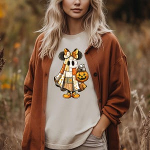 May include: A beige t-shirt featuring a cartoon ghost character with a patchwork design, black mouse ears, and a bow. The ghost is holding a jack-o'-lantern bucket. The character is wearing yellow shoes. The shirt is a Halloween-themed design.