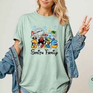 May include: A light green t-shirt with a colorful Disney-themed graphic. The graphic features the numbers "2026" with Disney characters and the words "Smith Family". The shirt is a Comfort Colors Bay.
