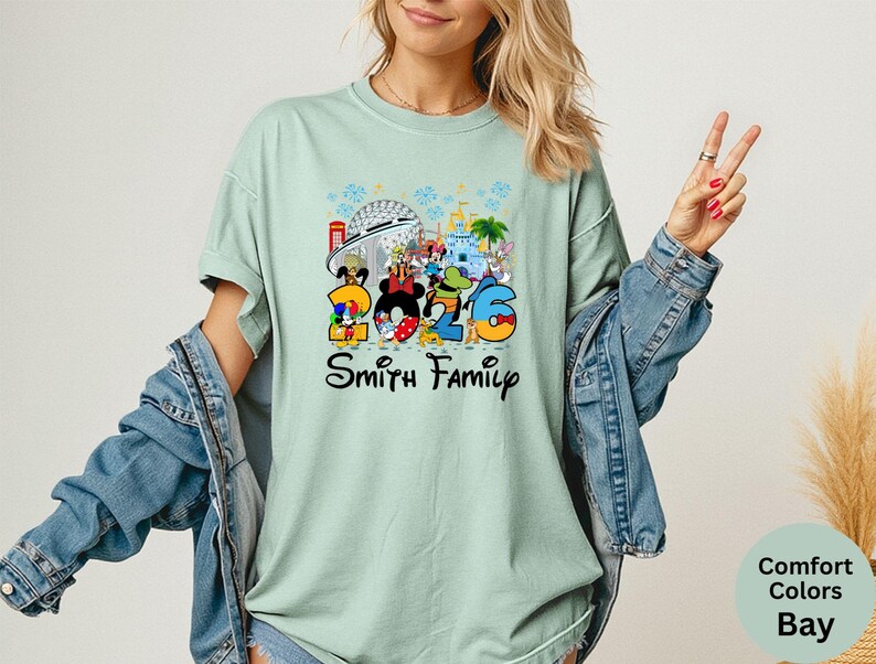 May include: A light green t-shirt with a colorful Disney-themed graphic. The graphic features the numbers "2026" with Disney characters and the words "Smith Family". The shirt is a Comfort Colors Bay.