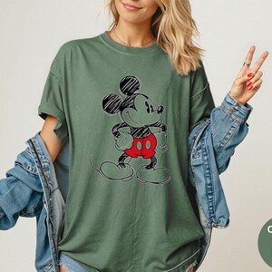 May include: A moss green t-shirt featuring a black and red Mickey Mouse graphic. The short-sleeved shirt has a relaxed fit and is paired with a denim jacket. The image also includes a small, round graphic with the words "Comfort Colors Moss".