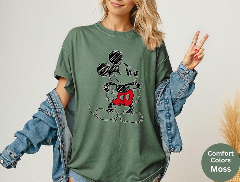 May include: A moss green t-shirt featuring a black and red Mickey Mouse graphic. The short-sleeved shirt has a relaxed fit and is paired with a denim jacket. The image also includes a small, round graphic with the words "Comfort Colors Moss".
