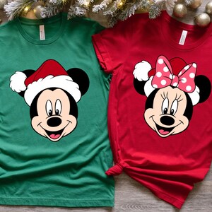 May include: Two Christmas themed t-shirts featuring Mickey Mouse and Minnie Mouse wearing Santa hats. The Mickey Mouse shirt is green and the Minnie Mouse shirt is red.
