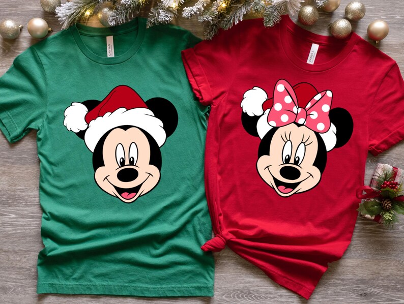 May include: Two Christmas themed t-shirts featuring Mickey Mouse and Minnie Mouse wearing Santa hats. The Mickey Mouse shirt is green and the Minnie Mouse shirt is red.