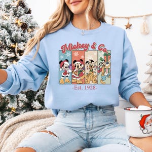May include: A light blue sweatshirt featuring a colorful graphic of Mickey Mouse, Minnie Mouse, Pluto, and Goofy in holiday attire. The words "Mickey & Co." are above the graphic, with "-Est. 1928-" below. The sweatshirt is a festive holiday item.