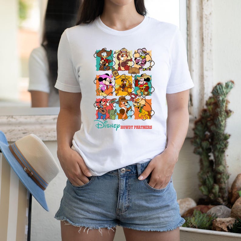 May include: White t-shirt featuring a colorful graphic of Disney characters in cowboy attire. The design includes Mickey Mouse, Minnie Mouse, Donald Duck, and Chip and Dale. The text "Disney Howdy Partners" is printed below the graphic.