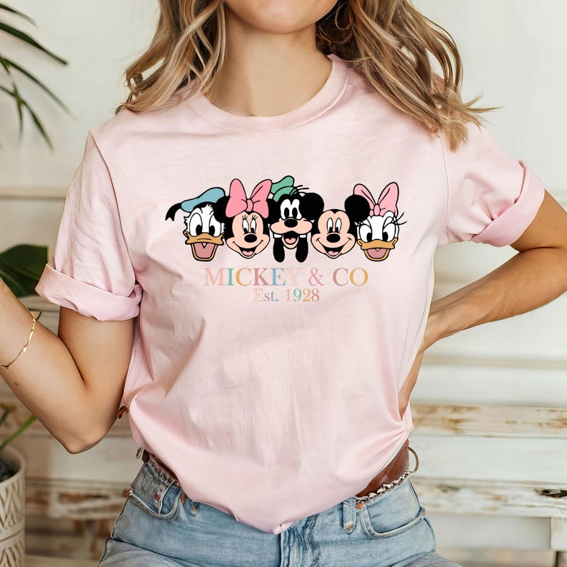 May include: Pink t-shirt with a graphic of Mickey Mouse and friends. The graphic includes Mickey Mouse, Minnie Mouse, Donald Duck, Goofy, and Pluto. The text below the graphic reads "Mickey & Co. Est. 1928".