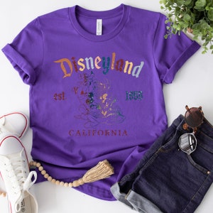 May include: Purple t-shirt with a vintage Disneyland graphic featuring Mickey Mouse and friends. The graphic includes the text "Disneyland est. 1955 California".