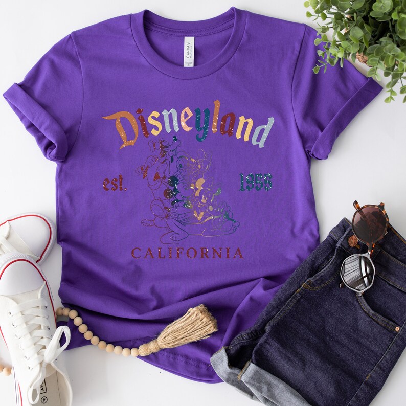 May include: Purple t-shirt with a vintage Disneyland graphic featuring Mickey Mouse and friends. The graphic includes the text "Disneyland est. 1955 California".