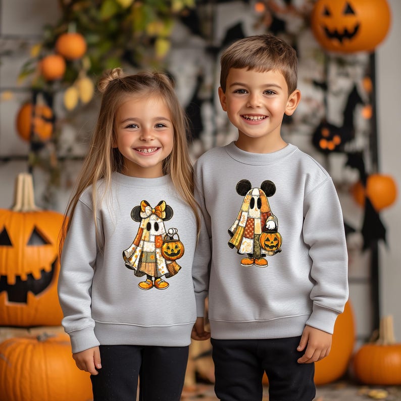 May include: Two children wearing matching gray sweatshirts with Halloween-themed designs. The sweatshirts feature a ghost character with Mickey Mouse ears, holding a jack-o'-lantern. The ghost is made of patchwork fabric. The children are smiling.