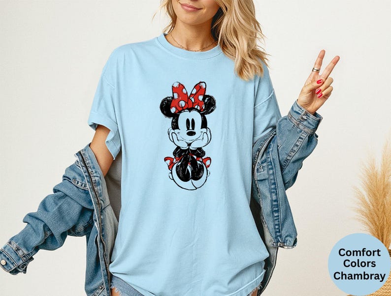 May include: Light blue t-shirt featuring a black and white Minnie Mouse graphic with a red polka dot bow. The shirt is part of the Comfort Colors Chambray collection.