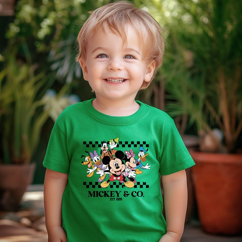 May include: A green crew neck t-shirt featuring a graphic of Mickey Mouse and friends. The design includes Mickey, Minnie, Donald Duck, Daisy Duck, Goofy, and Pluto. The text "Mickey & Co. EST 1928" is printed below the characters.