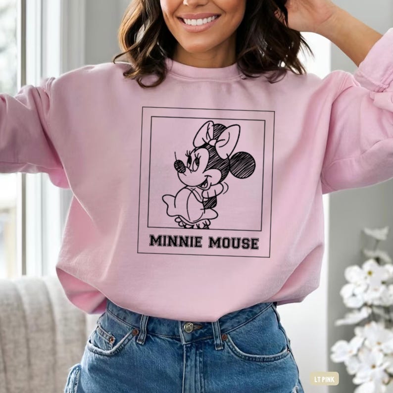 May include: A light pink sweatshirt featuring a black and white Minnie Mouse graphic within a square frame. The text "MINNIE MOUSE" is printed below the graphic in a bold, black font. The sweatshirt has long sleeves and a crew neck.